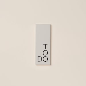 Puede incluir: Un bloc de notas rectangular gris claro con las palabras "TO DO" en negro, apiladas verticalmente. El bloc de notas está centrado sobre un fondo color crema, creando una estética minimalista.