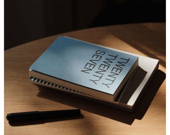 May include: A stack of two rectangular notebooks with a blue cover featuring the text TWENTY TWENTY SEVEN in black capital letters, accompanied by a black pen. The top notebook has a smooth blue surface, while the bottom one shows a striped edge.