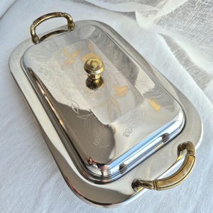 Peut inclure: Un plateau de service rectangulaire argenté avec un couvercle assorti, doté de deux poignées dorées sur les côtés et d'un bouton central doré. Le couvercle est orné de motifs floraux gravés et d'une petite étiquette rectangulaire dorée.