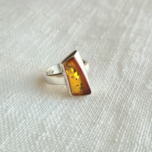 Può includere: Un anello in argento con un pezzo di ambra giallo oro traslucido di forma trapezoidale, lucidato e incastonato in una lunetta di metallo liscio. L'ambra presenta inclusioni naturali e una tonalità calda, montata su una fascia in argento massiccio.