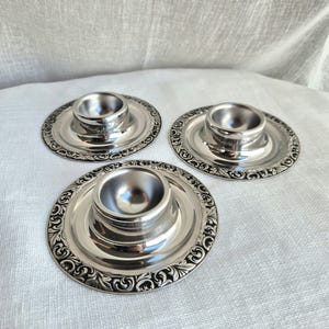 Puede incluir: Tres hueveras de metal plateado con acabado pulido. Cada huevera presenta un soporte circular central para un huevo, rodeado por un borde ancho y plano decorado con un intrincado patrón de volutas en relieve en el borde exterior.