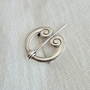 May include: A circular silver-toned brooch featuring a polished metallic finish. The design consists of a ring with two inward-curving spiral ends at the top, connected by a straight, pointed pin that extends diagonally across the center opening.
