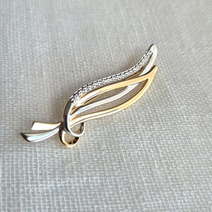 Peut inclure: Une broche en métal bicolore présentant un design incurvé en forme de plume. La pièce associe des finitions en métal doré et argenté poli, avec un bord orné d'une rangée de petites pierres claires et scintillantes le long de la courbe.