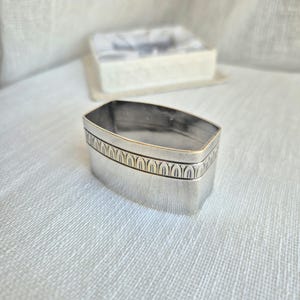 May include: A rectangular silver napkin ring with rounded corners, featuring a horizontal band of embossed vertical arch patterns around the upper section. The metal surface has a polished finish with visible signs of age and light tarnishing.