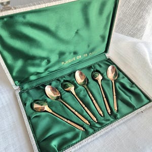 Pode incluir: Um conjunto de cinco colheres de metal dourado dispostas numa caixa de apresentação retangular com forro de cetim verde. O interior da tampa apresenta o texto em dourado PLAQUE OR 18 CARATS. A caixa tem um exterior texturizado claro.