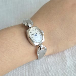 May include: A silver-toned analog wristwatch featuring an oval case with a white dial, black Arabic numerals, and blue hands. The watch face displays the text Kasper, 17 Rubis, and Antichoc. It has a metal link bracelet with engraved details.
