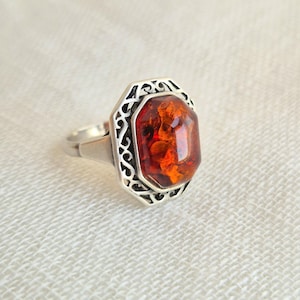 Peut inclure: Une bague en argent ornée d'une grande pierre d'ambre orange profond à facettes, sertie dans un cadre octogonal. La monture en métal présente un motif en filigrane ajouré complexe entourant la pierre centrale, sur un anneau lisse.
