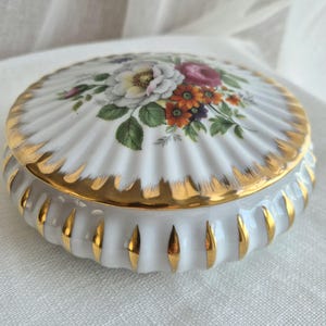 Può includere: Un portagioie rotondo in porcellana bianca con un design scanalato e accenti dipinti in oro lungo i bordi. Il coperchio presenta una colorata illustrazione di un bouquet floreale con fiori bianchi, rosa e arancioni circondati da foglie verdi.