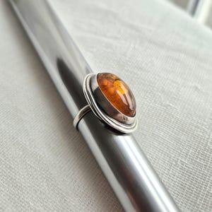 May include: A silver ring featuring a smooth, oval-shaped amber gemstone set in a polished metal bezel. The ring is positioned on a tapered metal mandrel, showcasing the warm, translucent orange tones of the amber and the metallic band.