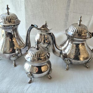 Peut inclure: Un service à thé en argent composé de quatre pièces : deux théières à couvercle nervuré, un sucrier couvert et un pot à lait. Chaque pièce présente une finition métallique polie, des gravures florales sur le corps et trois pieds en coquille.