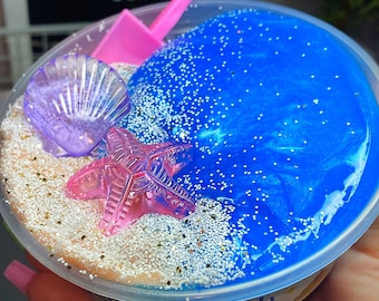 May include: A clear plastic container filled with blue and tan slime, decorated with a pink starfish, a purple seashell, and a pink shovel. The slime is sprinkled with glitter and has the text "Kgotyslimes.etsy.com".