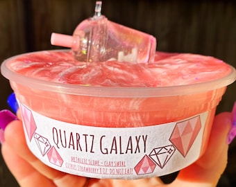 May include: A pink and white slime with a clear plastic charm shaped like a boba tea cup. The slime is in a clear plastic container with a label that reads "QUARTZ GALAXY METALLIC SLIME - CLAY SWIRL CITRUS STRAWBERRY 8 OZ DO NOT EAT!"