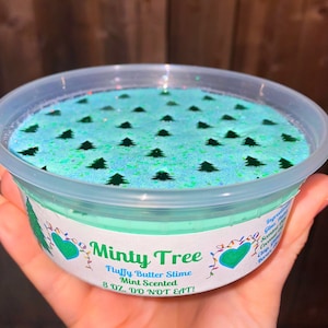 May include: A clear plastic container filled with light blue slime with green glitter and a pattern of green Christmas trees. The container has a white label that reads "Minty Tree Fluffy Butter Slime Mint Scented 8 OZ. DO NOT EAT!"