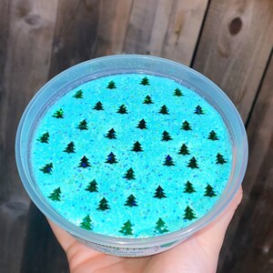 May include: A clear plastic container filled with light blue slime with green glitter and a pattern of green Christmas trees. The container has a white label that says "Fizzy Butter Slime" and "Hand Scented".