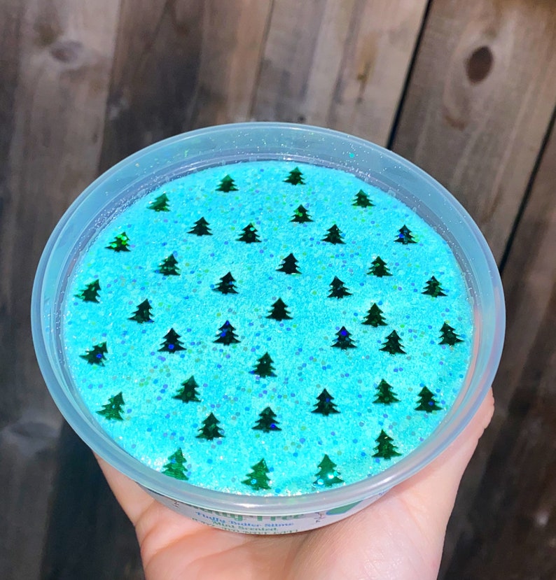 May include: A clear plastic container filled with light blue slime with green glitter and a pattern of green Christmas trees. The container has a white label that says "Fizzy Butter Slime" and "Hand Scented".