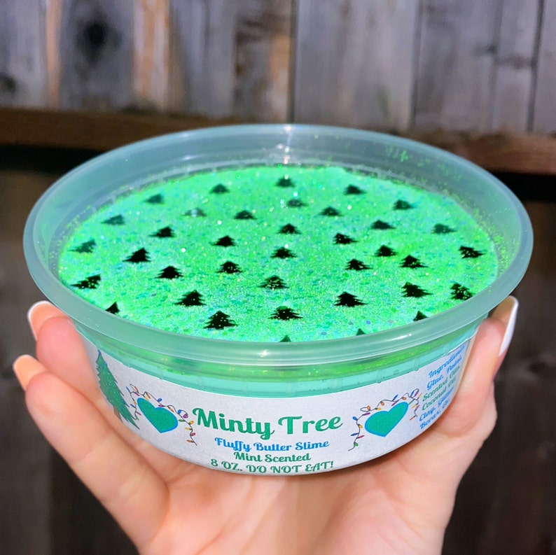 May include: A clear plastic container filled with green and black glitter slime. The slime is decorated with black silhouettes of Christmas trees. The container has a white label that reads "Minty Tree Fluffy Butter Slime Mint Scented 8 oz. DO NOT EAT!"