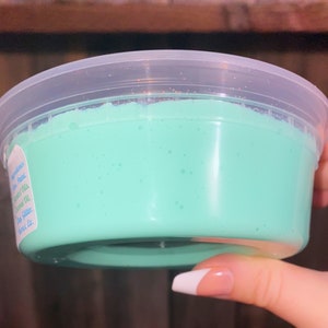May include: A clear plastic container filled with light green slime. The slime has a smooth, glossy texture and is slightly translucent.
