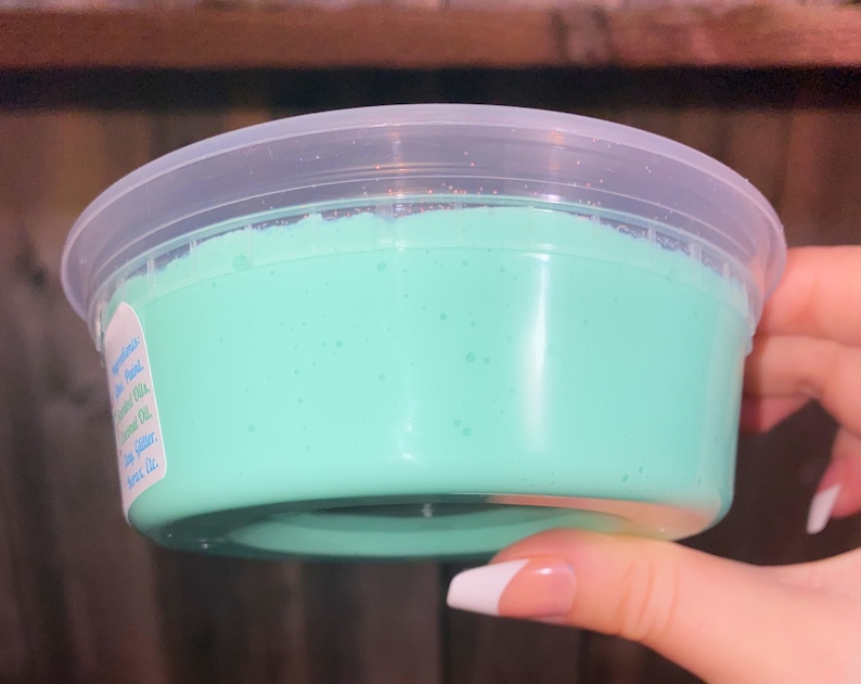 May include: A clear plastic container filled with light green slime. The slime has a smooth, glossy texture and is slightly translucent.