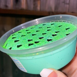 May include: A clear plastic container filled with light green slime with black glitter and black tree-shaped confetti on top.