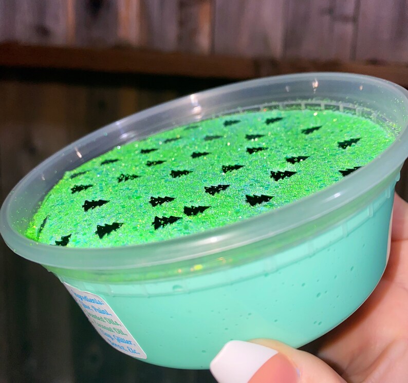 May include: A clear plastic container filled with light green slime with black glitter and black tree-shaped confetti on top.