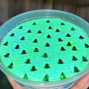 May include: A green and blue slime with a mint scent. The slime is in a clear plastic container with a white lid. The slime is decorated with small green glitter and green Christmas tree shapes.