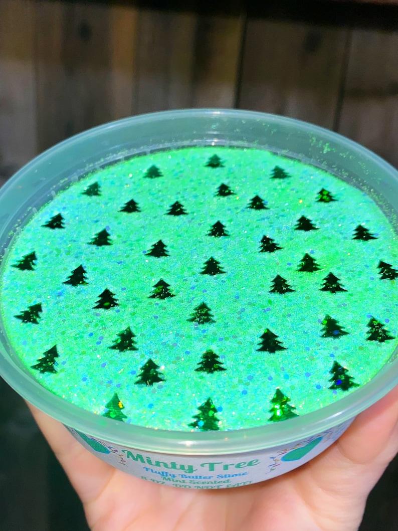 May include: A green and blue slime with a mint scent. The slime is in a clear plastic container with a white lid. The slime is decorated with small green glitter and green Christmas tree shapes.