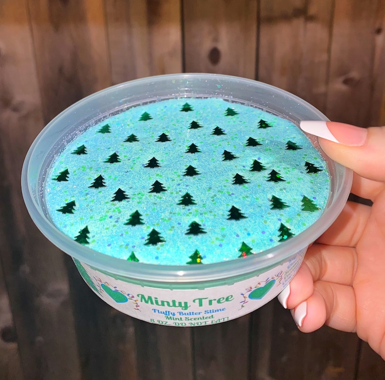 May include: A clear plastic container filled with light blue fluffy slime with green glitter and green Christmas tree shapes. The container has a green label that reads "Minty Tree Fluffy Butter Slime Mint Scented 8 oz. DO NOT EAT!"