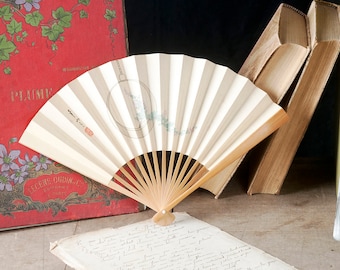 Op de afbeelding: Een rood hardback boek met een bloemenontwerp en de titel "Piéces Choisies" op de omslag. Een witte en beige handwaaier is open en rust op het boek. Een zwart-witfoto van twee kinderen is in een gouden lijst. De foto toont een jongen en een meisje die een fiets vasthouden.