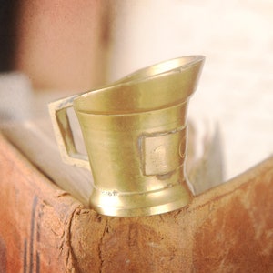 May include: A small, gold-colored metal cup with a handle, resting on the corner of an open, antique book. The book has a worn, brown leather cover. The background is blurred, suggesting other books and aged paper.