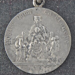 May include: Silver pendant featuring a religious image of the Virgin Mary with the baby Jesus. The inscription reads "Notre Dame La Blanche".