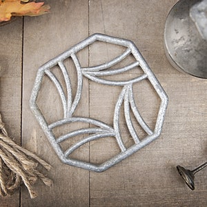 Może przedstawiać: Srebrny metalowy podkładka pod gorące z geometrycznym wzorem. Podkładka ma kształt sześciokąta i posiada ozdobny wzór z zakrzywionych linii.