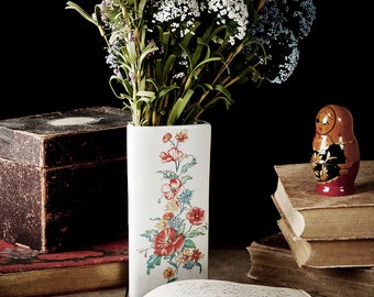 May include: A still life featuring a vintage glass spray bottle, a wooden box, a stack of old books, a vase with a floral pattern filled with purple flowers, and a small wooden nesting doll.