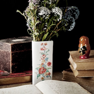 May include: A still life featuring a vintage glass spray bottle, a wooden box, a stack of old books, a vase with a floral pattern filled with purple flowers, and a small wooden nesting doll.