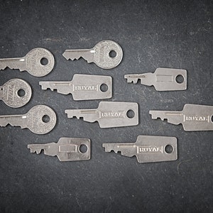 Può includere: Una collezione di dieci chiavi d'argento, alcune con la scritta "ROYAL" incisa su di esse. Le chiavi sono disposte a semicerchio su una superficie grigio scuro.