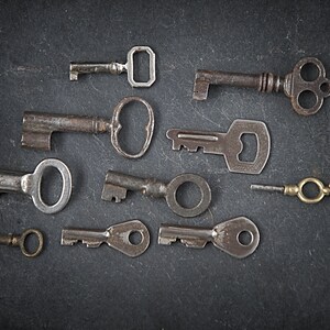 Puede incluir: Una colección de nueve llaves antiguas, algunas con diseños intrincados, dispuestas sobre una superficie gris oscuro. Las llaves son de metal y tienen un aspecto desgastado y envejecido.
