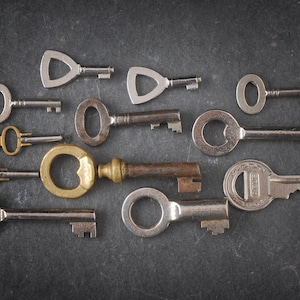 Puede incluir: Una colección de llaves vintage con diferentes diseños y acabados, incluyendo latón, plata y níquel. Las llaves están dispuestas sobre una superficie gris oscuro.