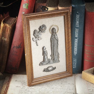 May include: A framed picture with a religious theme. The picture features a silver statue of the Virgin Mary with a rosary, a silver statue of a kneeling girl, and two silver roses.