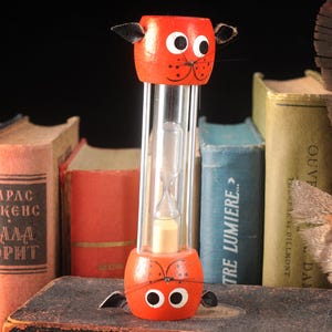 May include: A whimsical orange cat-shaped hourglass with a clear glass center, set against a backdrop of vintage books and a feather. The hourglass features black ears, eyes, and whiskers. The timer is a decorative item.