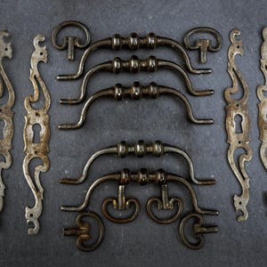 May include: An assortment of antique metal hardware, including ornate handles and keyhole plates, arranged on a dark gray surface. The hardware features a distressed, aged finish, suggesting vintage or rustic appeal. Ideal for furniture restoration.