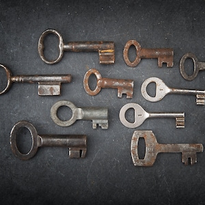 Puede incluir: Una colección de diez llaves antiguas de diferentes tamaños y formas, todas con una pátina oxidada, dispuestas sobre una superficie gris oscura.