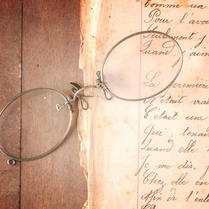 Puede incluir: Gafas antiguas plateadas con lentes ovaladas sobre un documento manuscrito antiguo. El documento tiene escritura cursiva y está envejecido. Las gafas están colocadas sobre una superficie de madera.