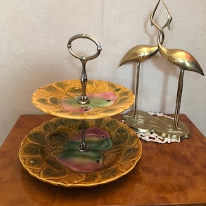 Peut inclure: Un présentoir à deux étages en céramique avec un motif de poires. Le présentoir a une poignée en métal et est décoré de poires vertes et roses sur un fond brun. Le présentoir est sur une table en bois à côté d'une sculpture en laiton de deux oiseaux.