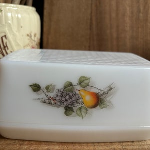 Peut inclure: Un beurrier en céramique blanche avec un couvercle surélevé. Le couvercle présente un motif peint à la main d'une poire et de raisins sur une vigne.