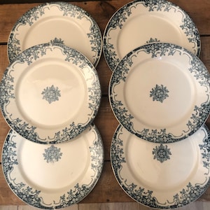 May include: Six white ceramic plates with a blue floral design around the rim and a central emblem. The plates are arranged on a wooden surface, showcasing their decorative pattern and vintage aesthetic.