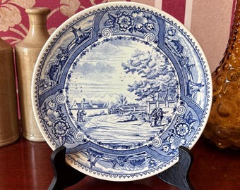 May include: A blue and white porcelain plate with a scenic design of a bridge and trees. The plate is on a black stand and has a white background with blue floral accents.