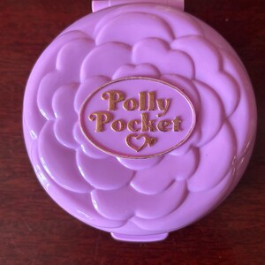 Op de afbeelding: Paarse plastic Polly Pocket speelgoed met een bloemmotief en gouden letters die "Polly Pocket" lezen met een hartje.