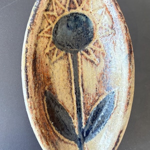 Peut inclure: Un plateau en céramique de forme ovale avec un émail beige et brun moucheté. Le centre présente un motif gravé à la main d'une fleur unique avec une tige, des feuilles et un cœur bleu foncé, entouré d'un motif en rayons de soleil gravé.