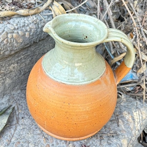 Peut inclure: Une cruche en céramique avec un dessus vert clair et un fond orange moucheté. La cruche a un corps arrondi et une anse.