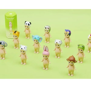 May include: A set of 12 small, nude-colored figurines wearing various animal hats. The hats include a panda, sloth, owl, chicken, koala, polar bear, bunny, frog, monkey, dalmatian, tiger, and elephant. The figurines are all standing on a green surface.