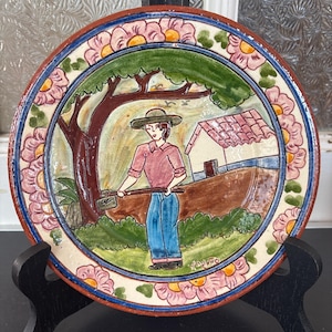 Peut inclure: Assiette en céramique peinte à la main avec une scène rurale. L'assiette présente une image centrale d'une personne avec une houe, une maison et un arbre. La bordure est ornée de fleurs roses et d'accents bleus. L'assiette est présentée sur un support noir.
